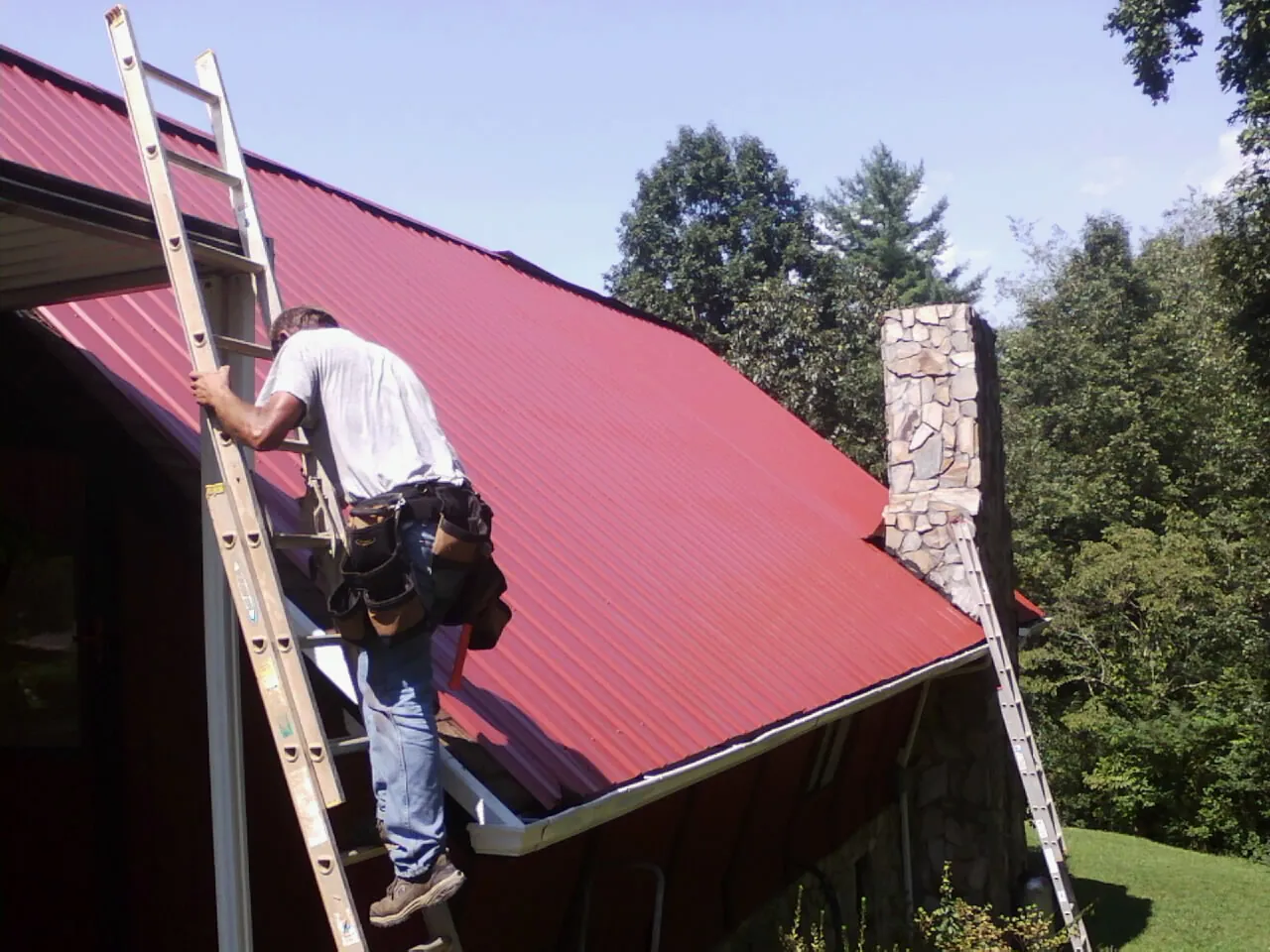 Roofer on a ladder beside a red metal roof and stone chimney — Soffit & Fascia Repair by Roofing Brothers in Lovilia